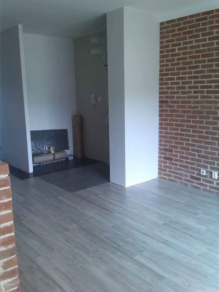 Wnętrze pokoju z panelami podłogowymi w odcieniach szarości, ceglaną ścianą i przygotowanym miejscem na kominek.