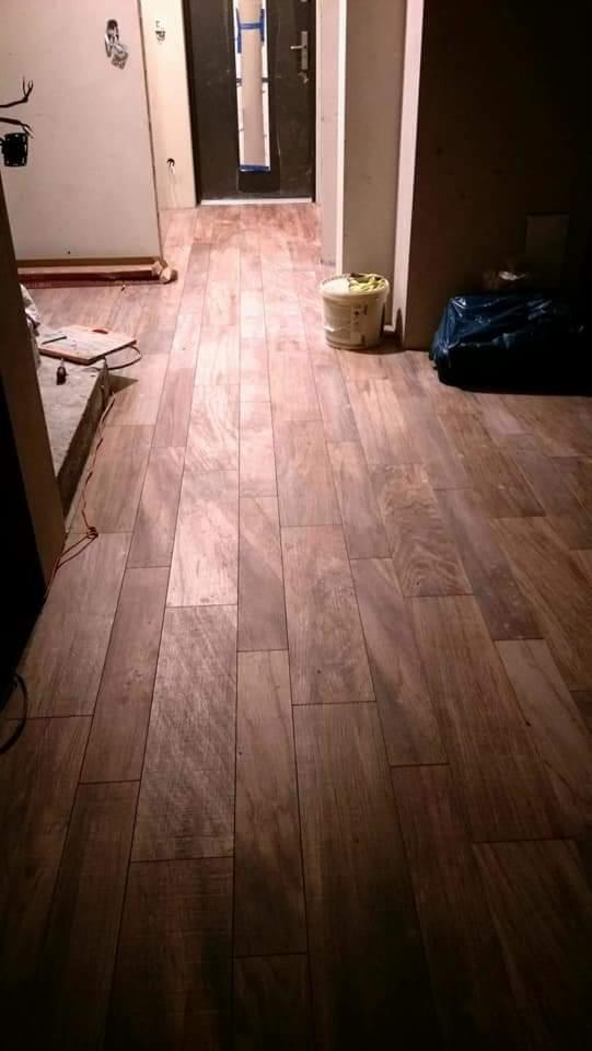 Ułożona podłoga z płytek imitujących drewno w korytarzu, widoczne wiadro i worek na śmieci, fragment ściany z instalacją elektryczną.