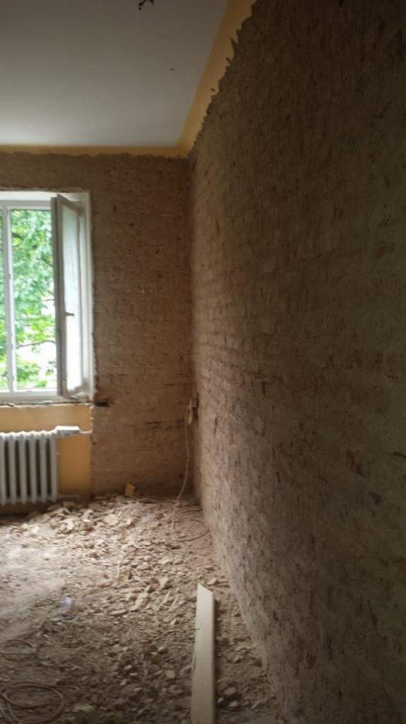 Pokój w trakcie remontu: odsłonięta cegła, gruz na podłodze, okno z widokiem na zieleń, stary grzejnik.