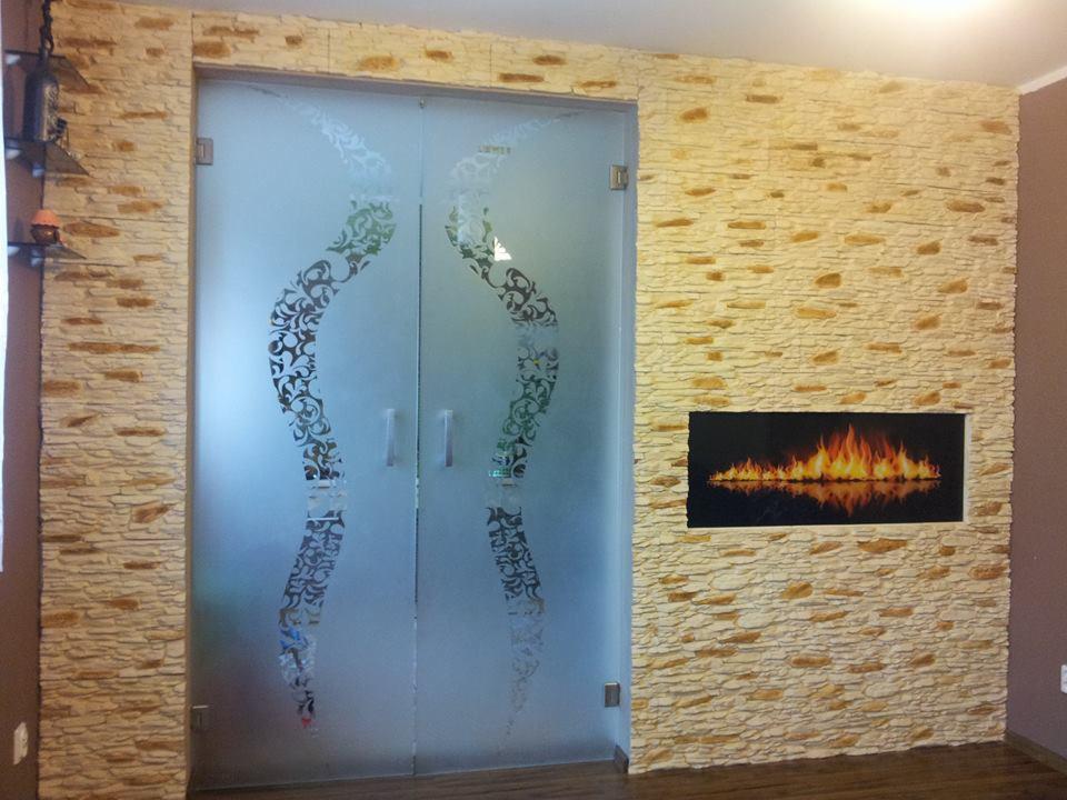 Szklane, przesuwne drzwi z ornamentem wkomponowane w ścianę z dekoracyjnej cegły, nad nimi półka z dekoracjami. Nowoczesny design.