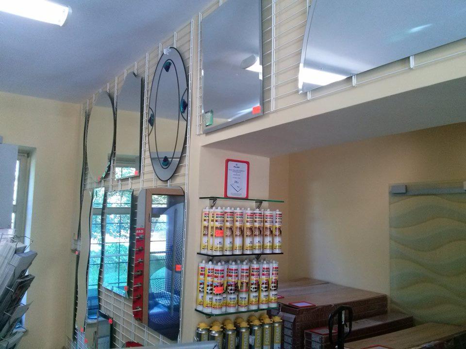 Ekspozycja luster o różnych kształtach i rozmiarach w sklepie szklarskim. Widoczne materiały montażowe na półkach i siatka ekspozycyjna.