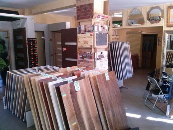 Ekspozycja paneli podłogowych w sklepie: różnorodne wzory drewna, od jasnych po ciemne, prezentowane w pionowych rzędach. W tle drzwi i lustra.