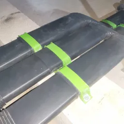 Rzędy czarnych, prostokątnych rur wentylacyjnych przymocowanych do podłoża za pomocą zielonych uchwytów, widoczne na tle betonowych elementów konstrukcyjnych.