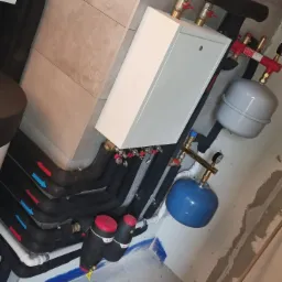 Nowoczesny system grzewczy z białym piecem, czarnymi rurami z oznaczeniami przepływu (czerwone i niebieskie strzałki), oraz niebieskim zbiornikiem wyrównawczym, zamontowany na tle surowej ściany.