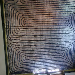 Rozłożona instalacja ogrzewania podłogowego z niebieskich rurek na srebrnej folii izolacyjnej, widok z góry, w tle okno z widokiem na zieleń.