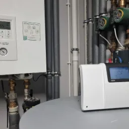 Nowoczesna instalacja grzewcza z pompą ciepła Panasonic Aquarea, widoczne rury, zawory i sterownik Tech na tle szarej ściany.