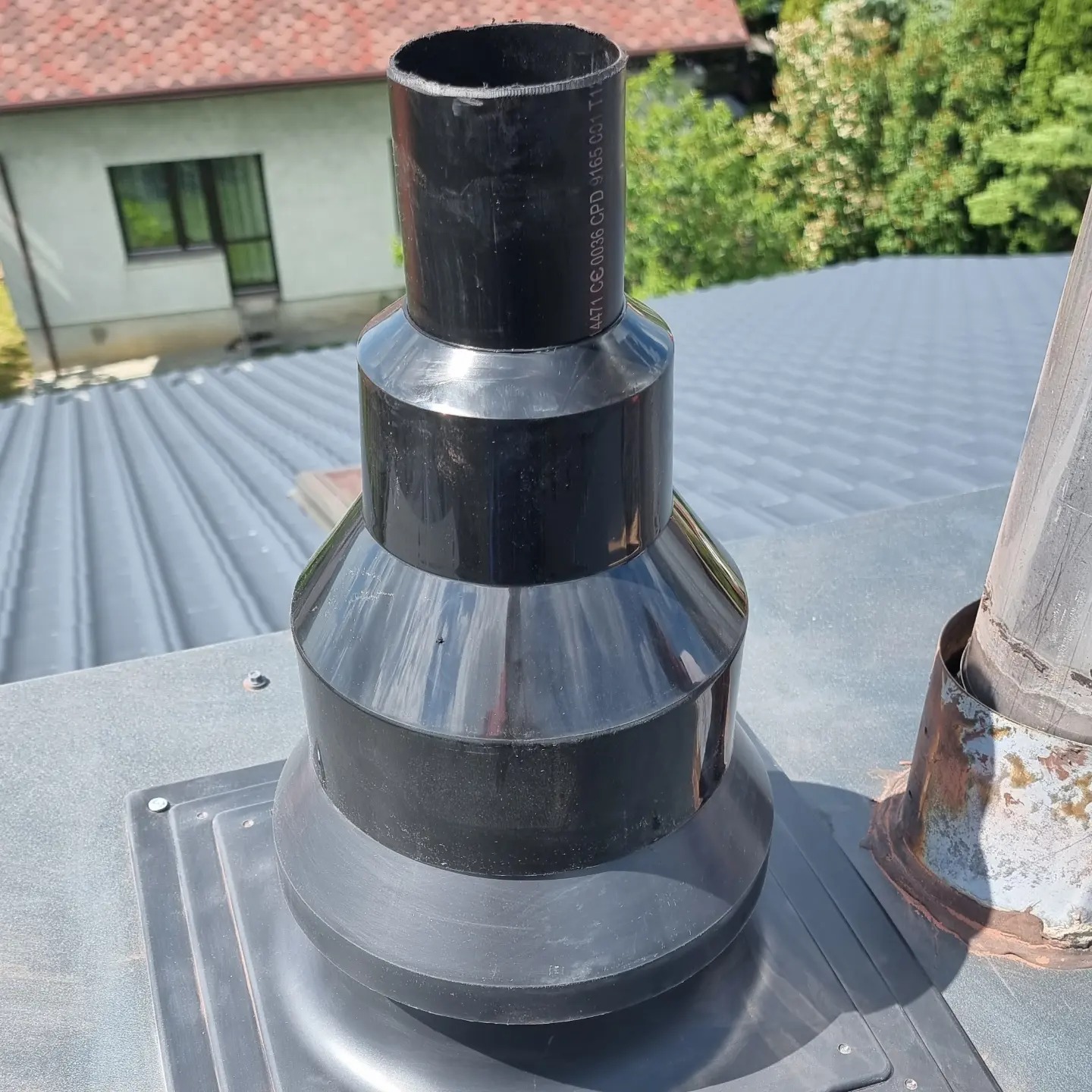 Czarny, błyszczący komin wentylacyjny z tworzywa sztucznego, zamontowany na szarej blasze trapezowej dachu, obok skorodowanego komina metalowego. Widoczne oznaczenia na rurze.