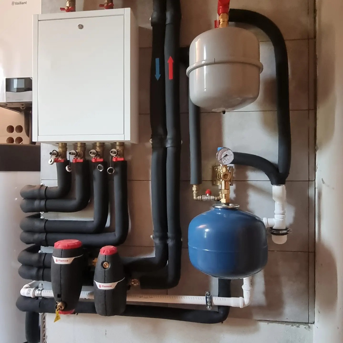 Złożona instalacja grzewcza z piecem Vaillant, czarnymi rurami izolowanymi, dwoma zbiornikami wyrównawczymi (szarym i niebieskim) oraz filtrami Flamco, zamontowana na ścianie z płytek.