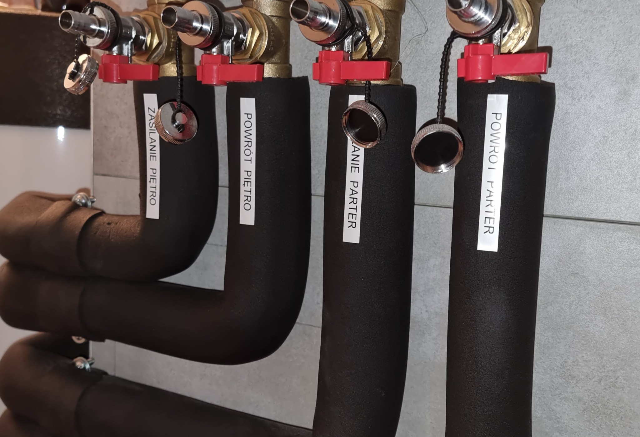 Cztery zaizolowane rury instalacji grzewczej z zaworami i etykietami 'Zasilanie Piętro', 'Powrót Piętro', 'Zasilanie Parter', 'Powrót Parter' na jasnoszarej ścianie.