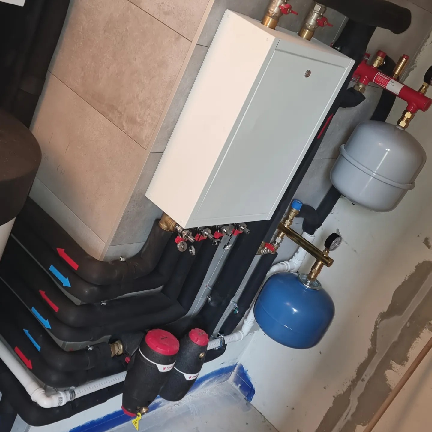 Nowoczesny system grzewczy z białym piecem, czarnymi rurami z oznaczeniami przepływu (czerwone i niebieskie strzałki), oraz niebieskim zbiornikiem wyrównawczym, zamontowany na tle surowej ściany.