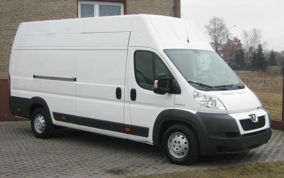 Biały, wysoki bus Peugeot na kostce brukowej, widok z boku i przodu. Pojazd z relingami na dachu, czarne elementy wykończenia. Tło: budynek i drzewa.