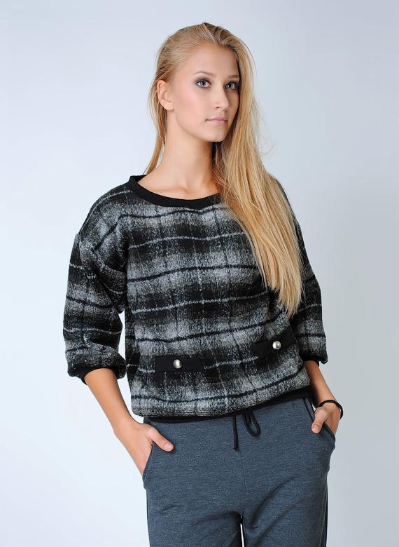 Blondwłosa kobieta prezentuje szaro-czarny sweter w kratę z trzema guzikami, zestawiony z szarymi spodniami dresowymi na białym tle.