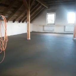 Nowa, gładka wylewka podłogowa w surowym wnętrzu poddasza, widoczne drewniane słupy i krokwie, przewód elektryczny zwisający z boku.