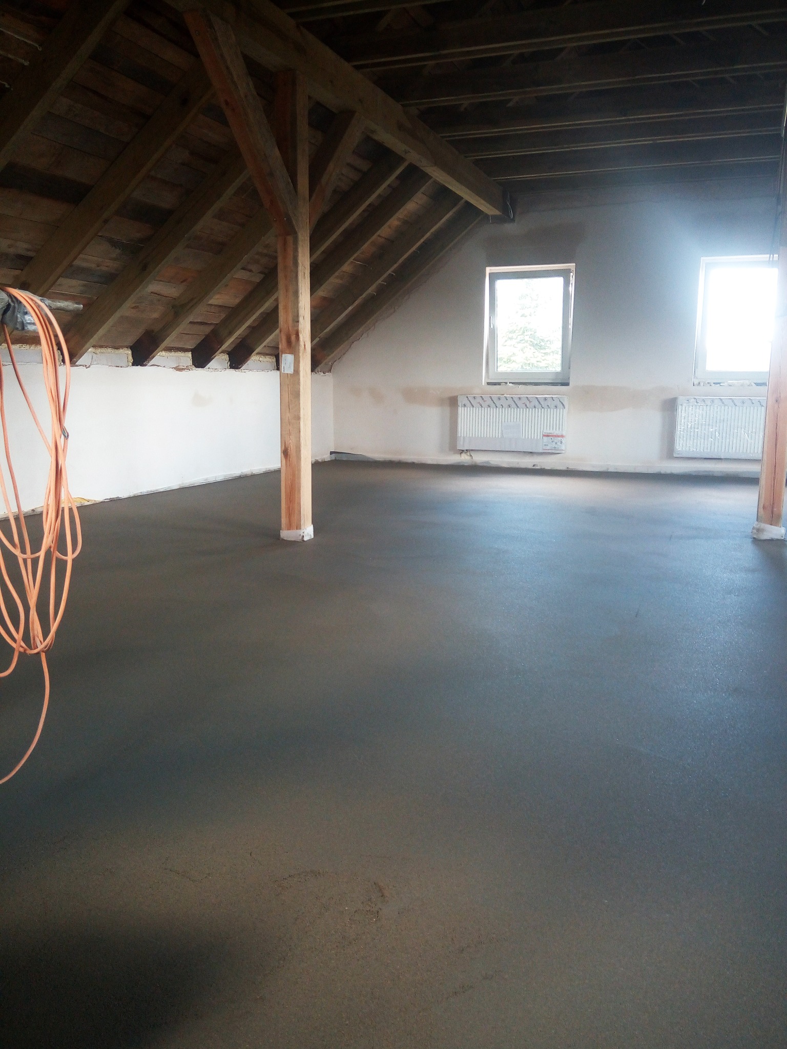 Nowa, gładka wylewka podłogowa w surowym wnętrzu poddasza, widoczne drewniane słupy i krokwie, przewód elektryczny zwisający z boku.