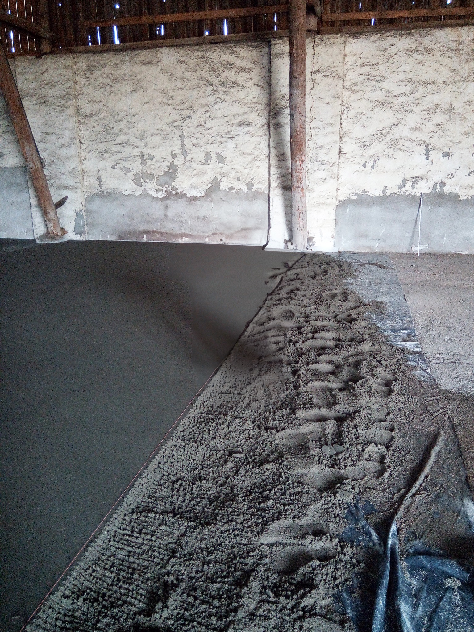 Wylewka podłogowa w trakcie wykonywania w surowym wnętrzu budynku gospodarczego, widoczne ślady stóp na świeżym materiale i fragment folii zabezpieczającej.