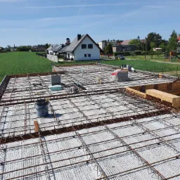 Zbrojenie płyty fundamentowej z widocznym szalunkiem i elementami izolacji termicznej, w tle zabudowa jednorodzinna i zielone pola.