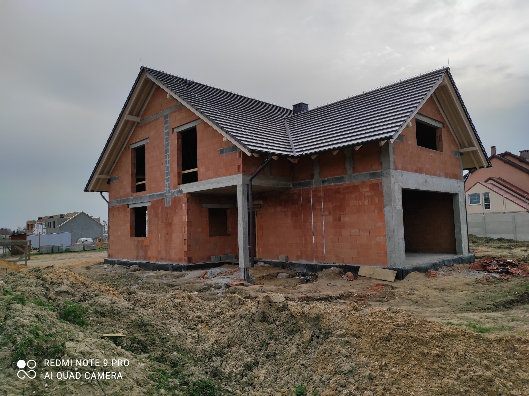 Surowy stan domu jednorodzinnego z czerwonej cegły z czarną dachówką, widoczne otwory okienne i betonowe słupy, ziemia wokół nierówna, w tle inne budynki.