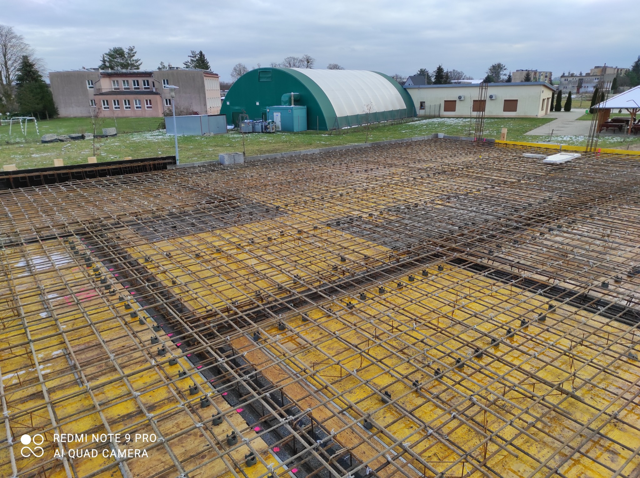 Zbrojenie płyty fundamentowej na żółtym szalunku, widoczne dystanse pod zbrojeniem oraz pionowe pręty zbrojeniowe wystające z gotowych otworów, w tle budynki i częściowo ośnieżony teren.