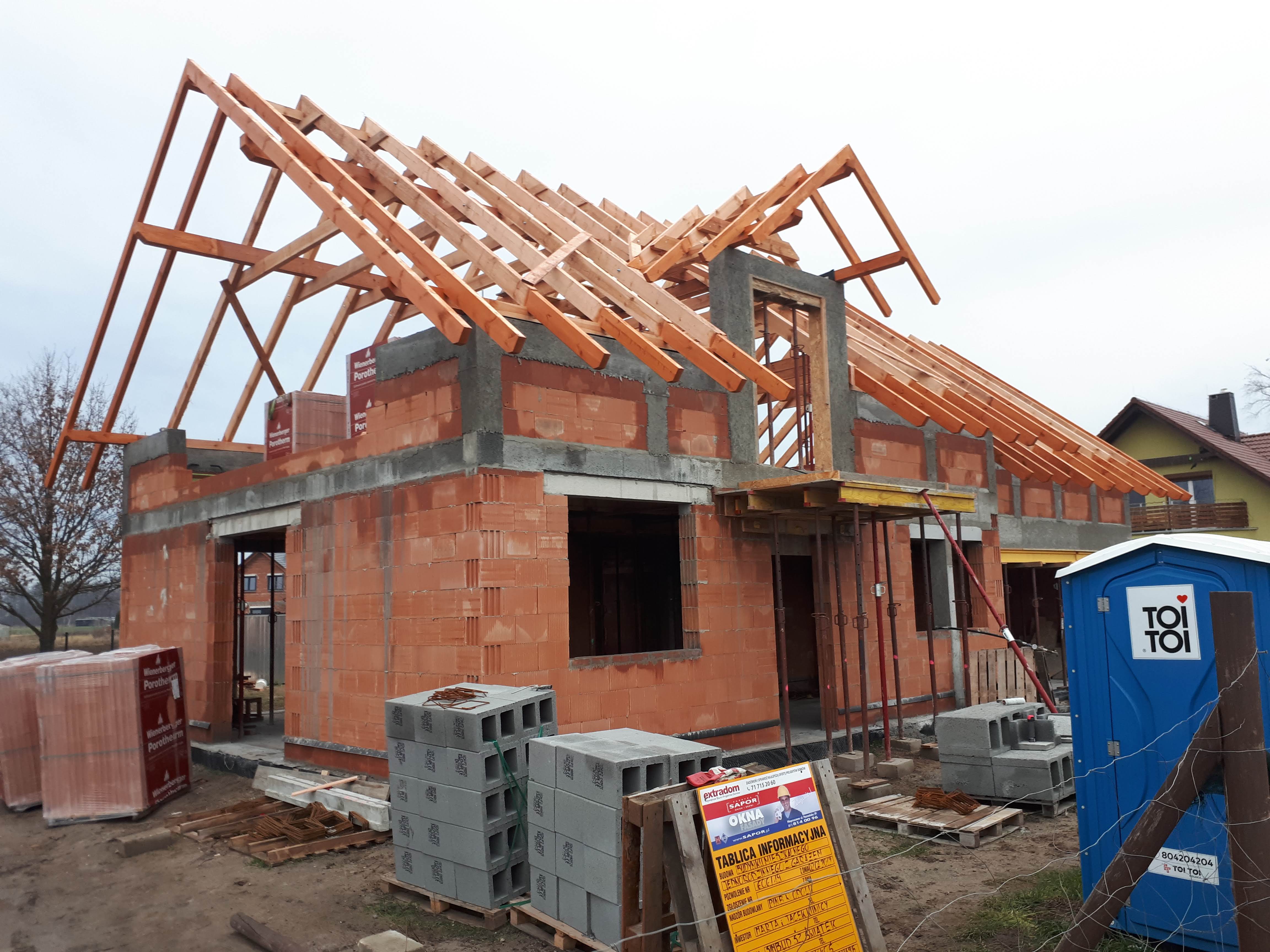 Budowa domu jednorodzinnego z czerwonej cegły: widoczne ściany, drewniana konstrukcja dachu w trakcie montażu, palety z bloczkami oraz tablica informacyjna budowy.