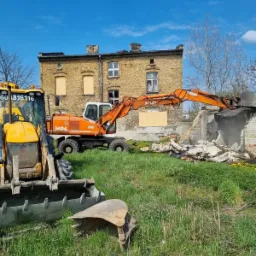 Pomarańczowa koparka Atlas 1404 w trakcie kruszenia muru przy żółtym budynku z zabitymi oknami, widoczna także żółta koparko-ładowarka na pierwszym planie, trawa i gruz na terenie budowy.