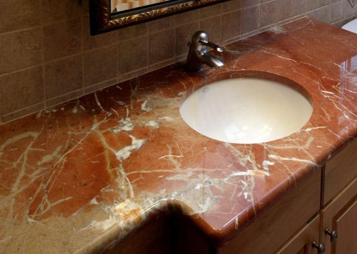 Błyszczący, czerwono-biały blat kamienny z umywalką i chromowaną baterią w łazience z brązowymi płytkami i lustrem w ozdobnej ramie.