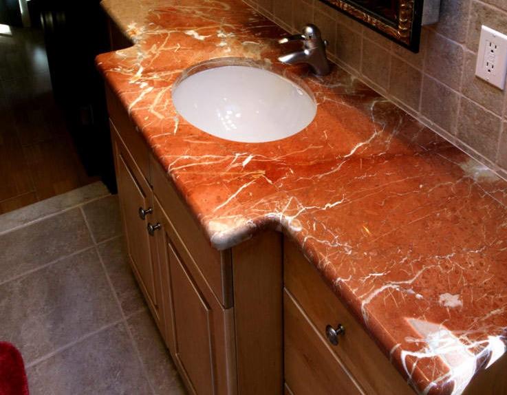 Czerwony blat kamienny z umywalką nablatową, zamontowany na drewnianej szafce w łazience. Widoczna bateria i fragment lustra.