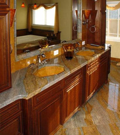 Łazienka z blatem kamiennym o unikalnym wzorze i drewnianymi szafkami. Umywalki w kolorze blatu, lustro z widokiem na okno. Podłoga wyłożona płytkami imitującymi kamień.