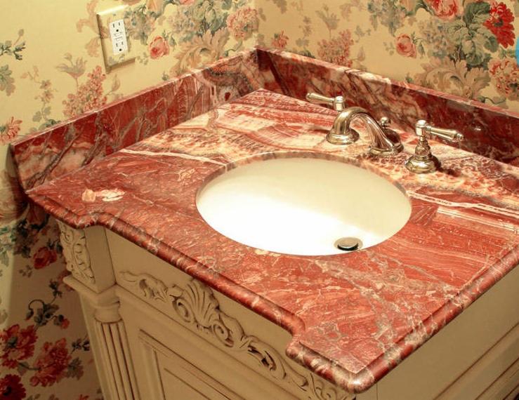 Blat łazienkowy z czerwonego marmuru z umywalką i ozdobną szafką, na tle ściany w kwiatowe wzory, oświetlony od góry.