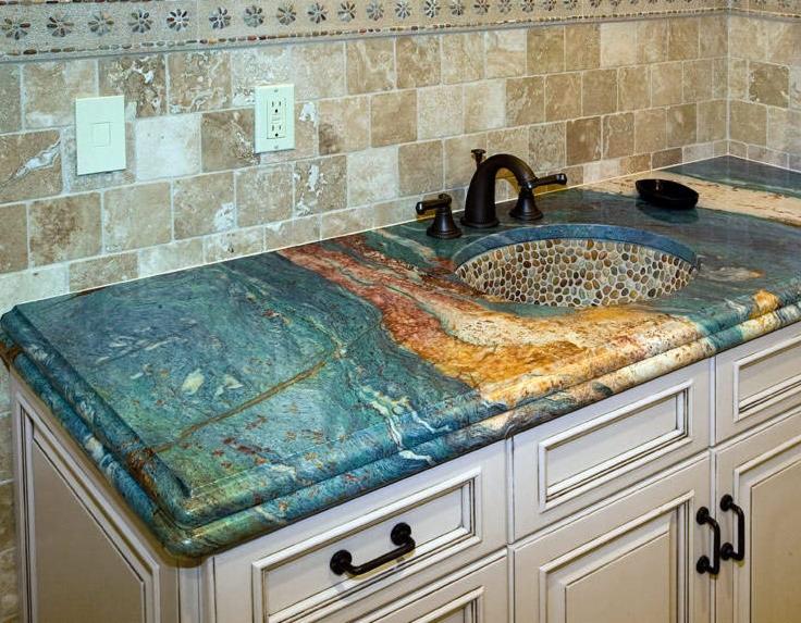 Błękitno-złoty blat kamienny z oryginalną umywalką z kamieni rzecznych, zamontowany na białej szafce w łazience z beżowymi płytkami. Nietypowy wzór blatu.