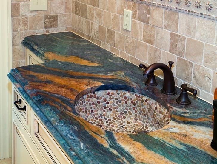 Unikalny blat kamienny z wbudowaną umywalką z kamiennych otoczaków, wykończony w odcieniach niebieskiego, brązu i złota, z ciemną armaturą na tle beżowej mozaiki.