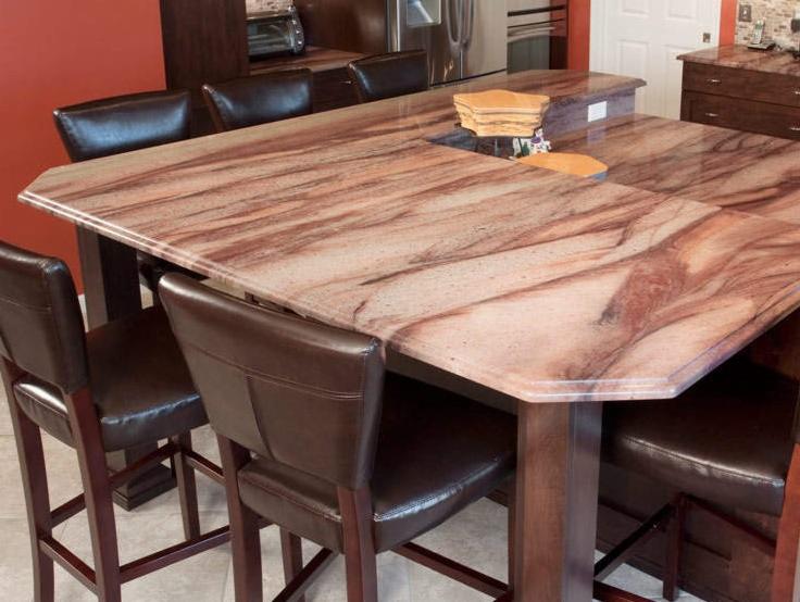 Rozległy blat z kamienia o unikalnym wzorze w kuchni w Ostrołęce, otoczony eleganckimi, brązowymi krzesłami barowymi. Ciepłe oświetlenie podkreśla detale.