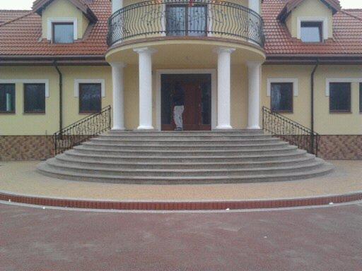 Eleganckie schody kamienne z balustradami prowadzące do wejścia budynku z kolumnami i balkonem. Widok z dołu ukazuje symetryczny układ i detale wykończenia.