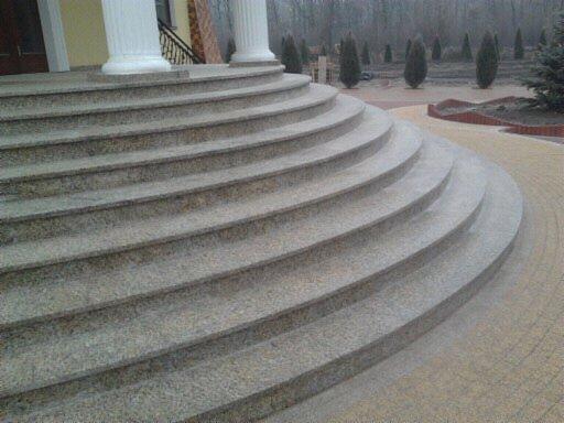 Granitowe schody zewnętrzne z kolumnami, prowadzące do budynku. Widoczne zaokrąglone stopnie i fragment chodnika z kostki brukowej. Tło stanowią drzewa.