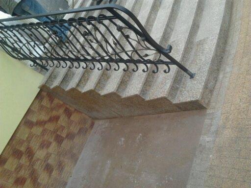 Kamienne schody zewnętrzne z ozdobną, kutą balustradą, widziane pod kątem. Ściana z żółtym odcieniem w tle. Widoczne detale wykończenia.