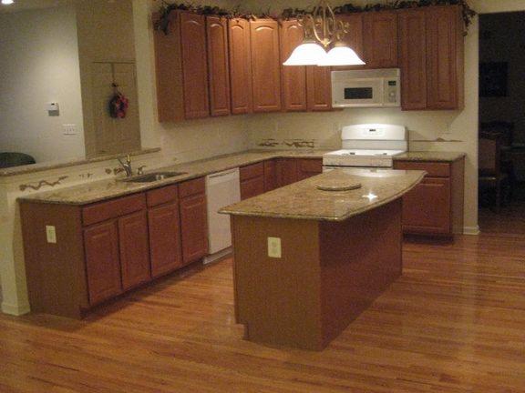 Kuchnia z drewnianymi szafkami i blatem z jasnego kamienia. Wyspa kuchenna z blatem o nieregularnym kształcie. Parkiet na podłodze. Dekoracja nad szafkami.