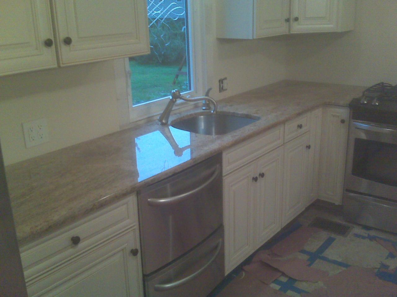 Jasny blat kamienny w kuchni z oknem, zlewozmywakiem i armaturą. Widoczne białe szafki, sprzęt AGD i zabezpieczona podłoga. Blat odbija światło.