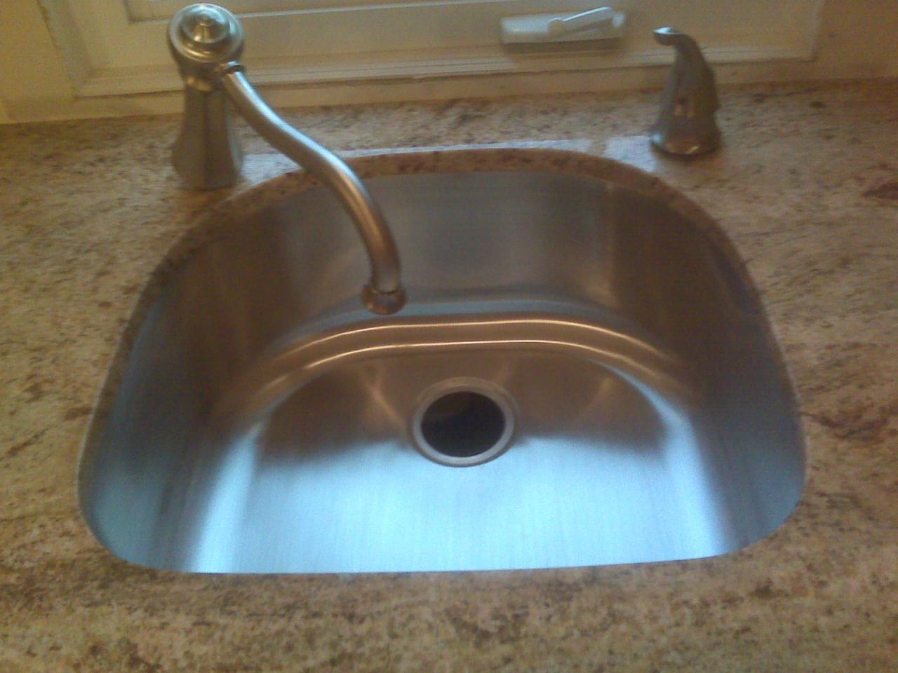 Błyszczący, metalowy zlew wpuszczony w blat kamienny o nakrapianej fakturze, z widoczną baterią i dozownikiem, umieszczony pod oknem.