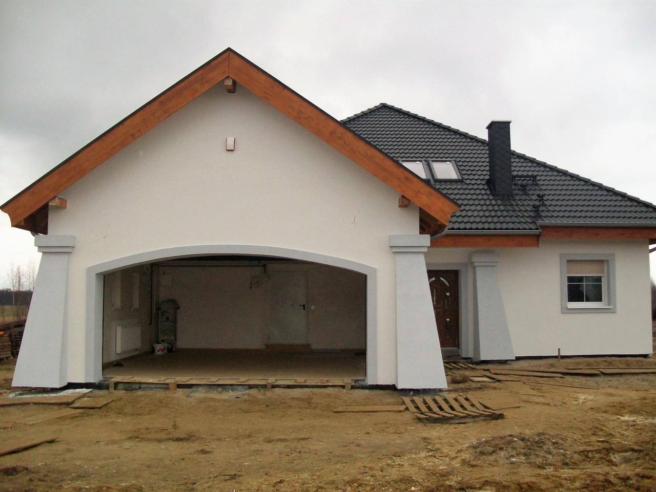 Nowoczesny dom jednorodzinny z garażem w trakcie wykończenia elewacji, widoczne elementy drewniane dachu i szara dachówka. Otoczenie placu budowy.