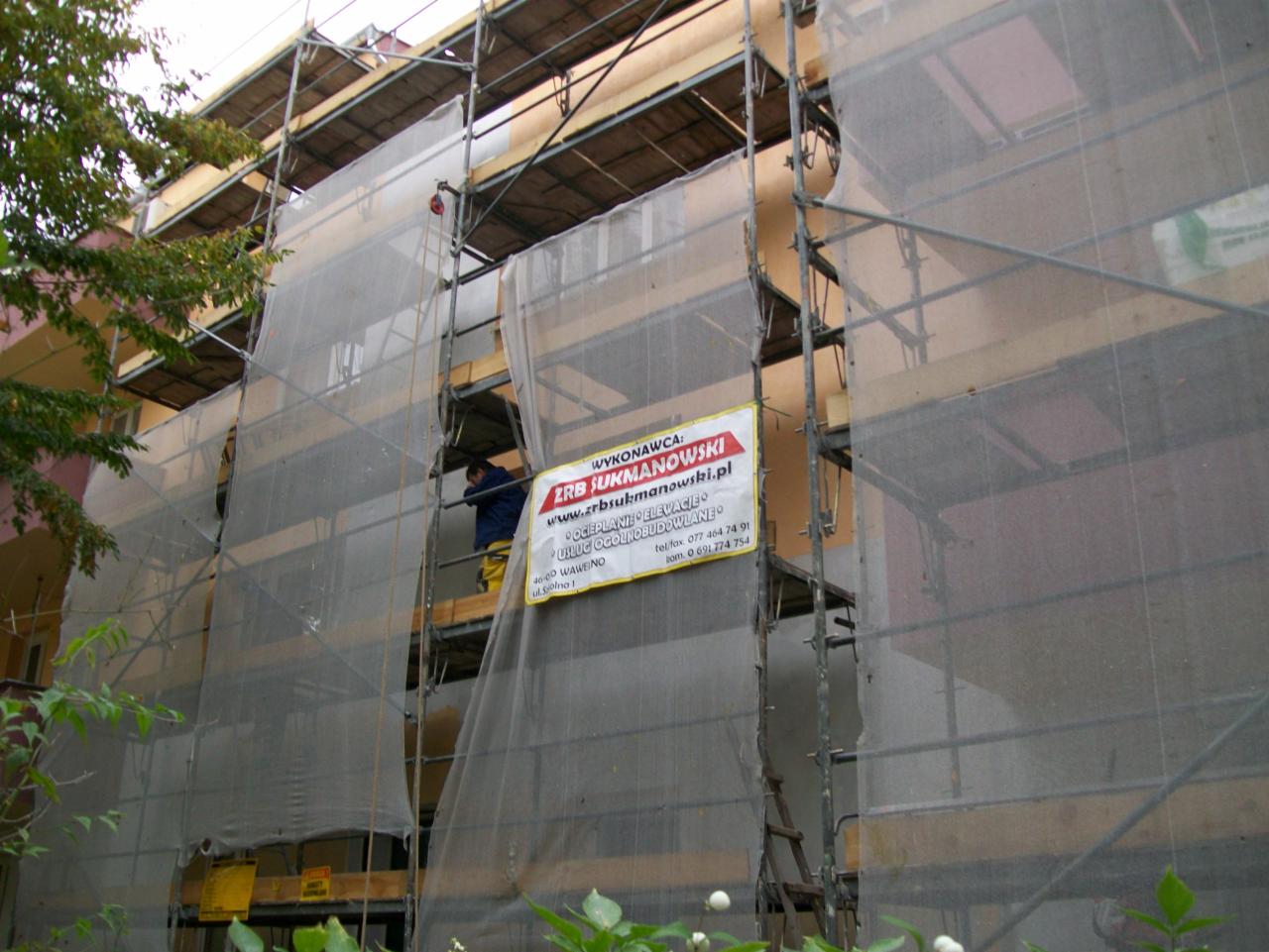 Budynek w rusztowaniach osłonięty siatką, widoczny pracownik w roboczym ubraniu. Na rusztowaniu baner firmy ZRB SUKMANOWSKI oferującej ocieplanie i elewacje.
