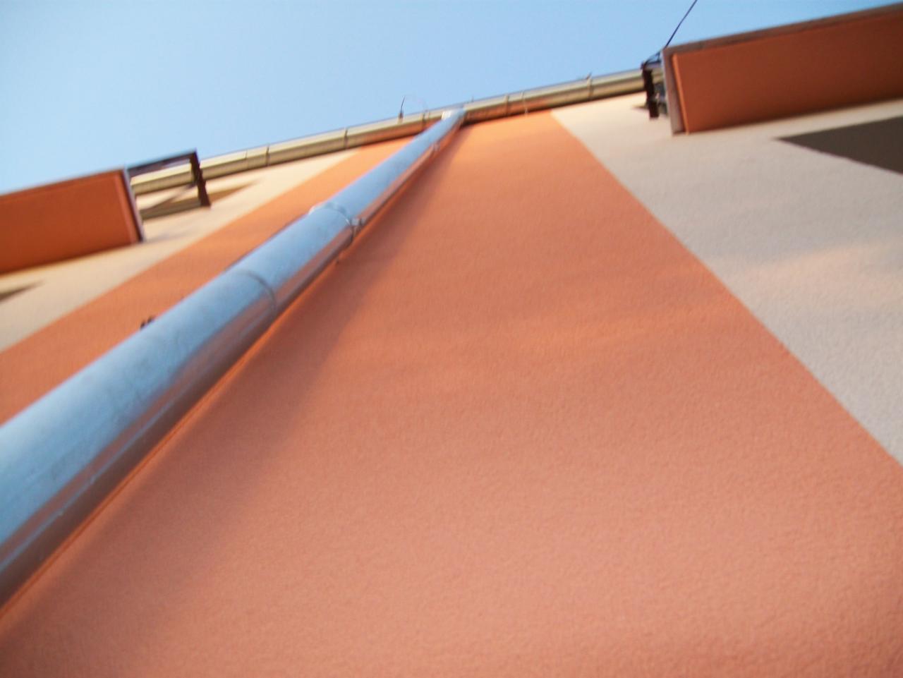 Nisko kątowe ujęcie pomalowanej elewacji w odcieniach pomarańczy i beżu z rynną. Widoczny fragment dachu i błękitne niebo. Charakterystyczny układ kolorów.