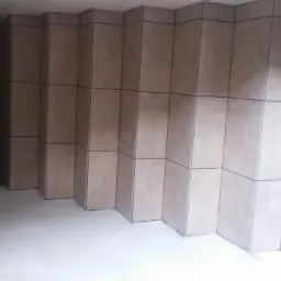 Wykończona ściana z ułożonych, beżowych, kwadratowych płytek w kształcie schodków. Na podłodze widać jasną wylewkę. Część ściany pomalowana na ciemny kolor.