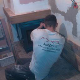 Joddom - Pracownik w koszulce firmowej JodDom montuje wkład kominkowy w trakcie prac wykończeniowych, widoczne niedokończone ściany i fragment podłogi z płytek.