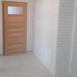 Joddom - Jasne wnętrze po remoncie: drewniane drzwi z szybą, ściana z białej cegły i podłoga z płytek imitujących drewno.