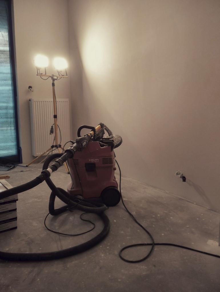 Przygotowanie do szlifowania ściany: lampa halogenowa, odkurzacz przemysłowy Hilti i szlifierka na podłodze w pokoju przed malowaniem.