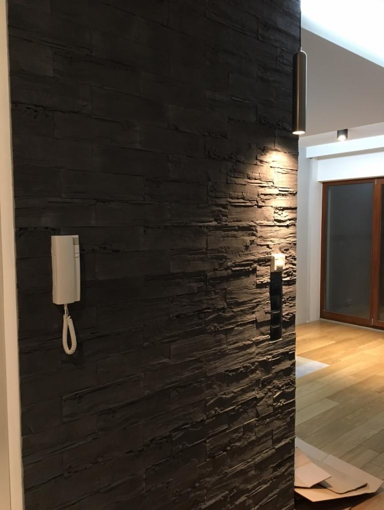 Ściana wyłożona ciemnym kamieniem dekoracyjnym z widocznym domofonem, włącznikiem światła i lampą wiszącą w minimalistycznym stylu, podłoga wyłożona parkietem.