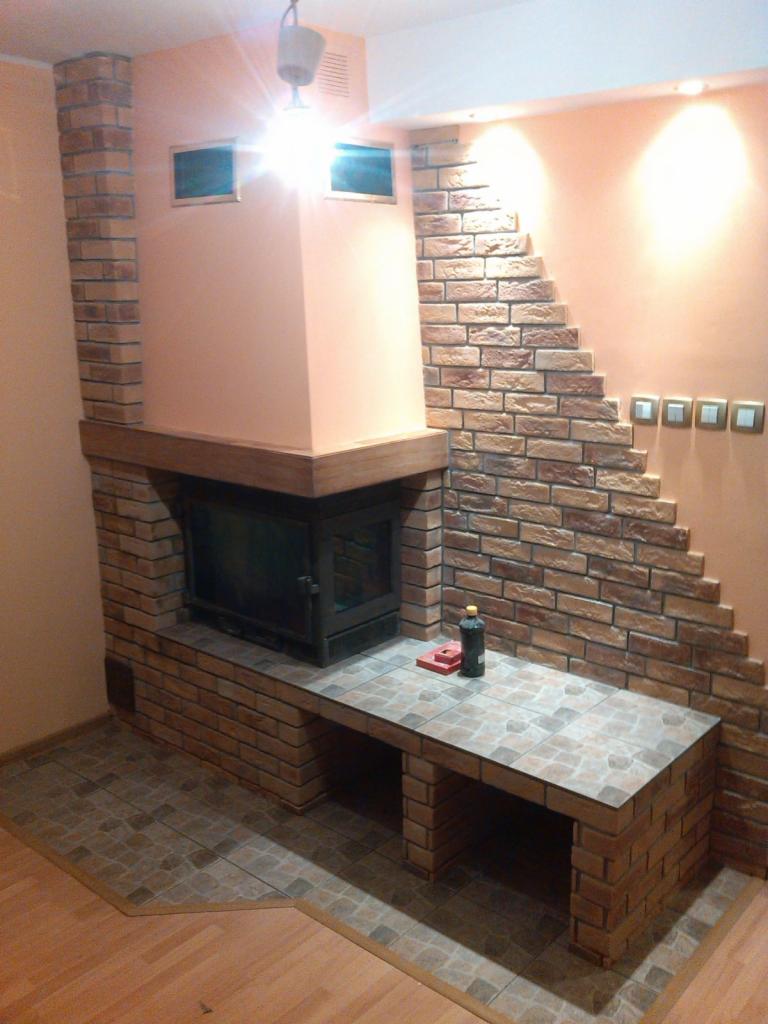 Wykończony kominek z cegły z półką z płytek, z butelką płynu do czyszczenia i czerwonym narzędziem na blacie, podświetlony przez dwa reflektory i lampę wiszącą.