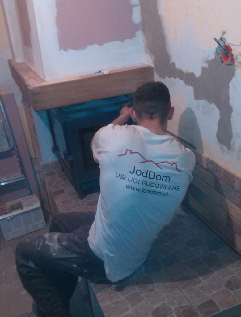 Pracownik w koszulce firmowej JodDom montuje wkład kominkowy w trakcie prac wykończeniowych, widoczne niedokończone ściany i fragment podłogi z płytek.