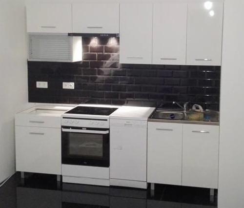 Biała kuchnia z czarnymi, błyszczącymi płytkami na ścianie, widok na zabudowę kuchenną z piekarnikiem, zlewem i szafkami, czarna podłoga z połyskiem.