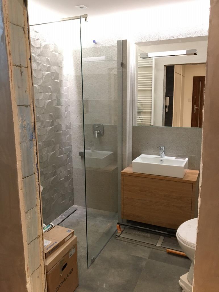 Półotwarta łazienka z kabiną prysznicową typu walk-in, umywalką nablatową na drewnianej szafce i szarą, wzorzystą ścianą w strefie prysznica, widoczne kartony z materiałami budowlanymi.