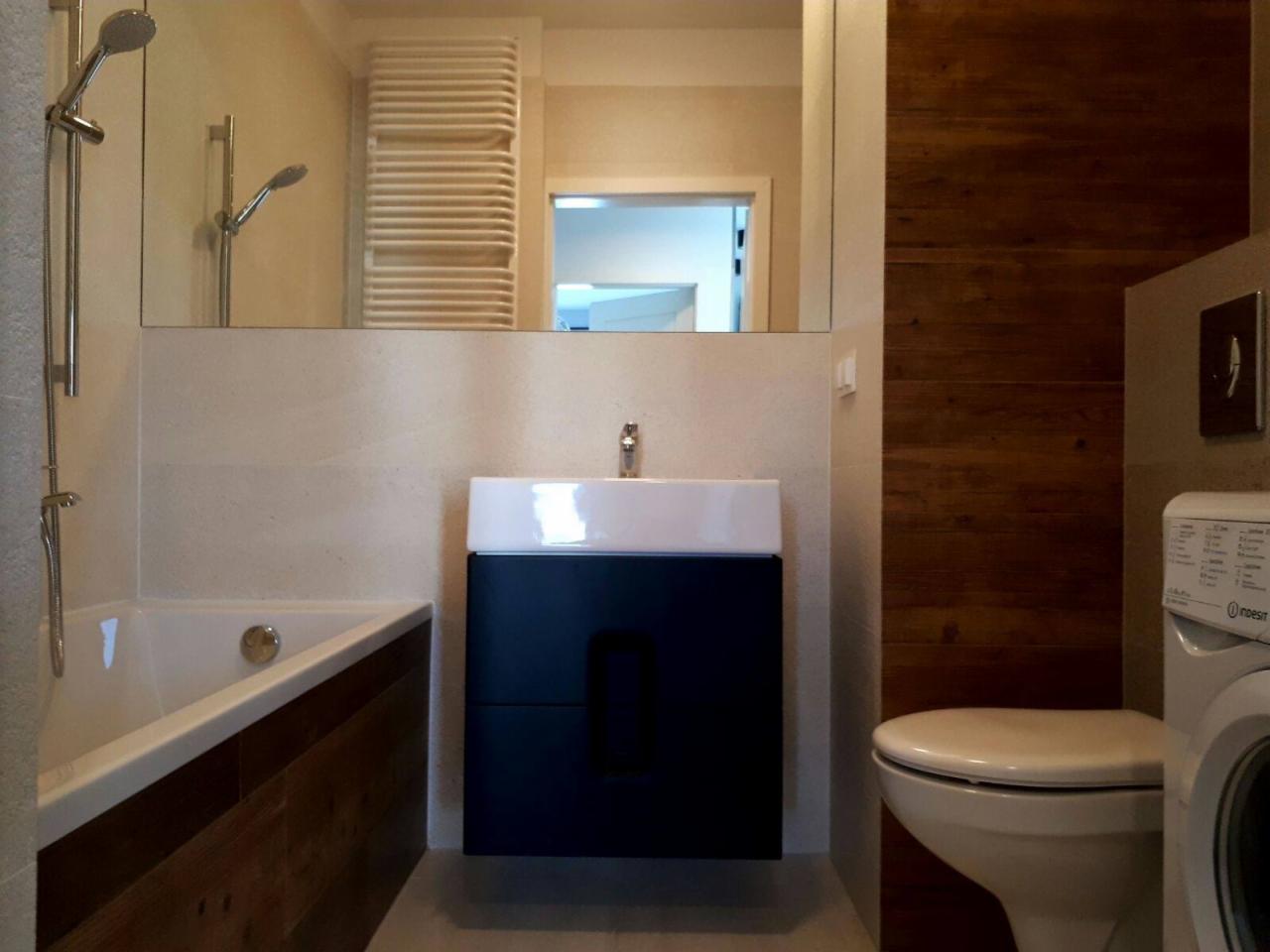 Nowoczesna łazienka z wanną obudowaną drewnem, umywalką na szafce w kolorze granatowym, toaletą i pralką, całość dopełniona drewnianą ścianą i dużym lustrem.